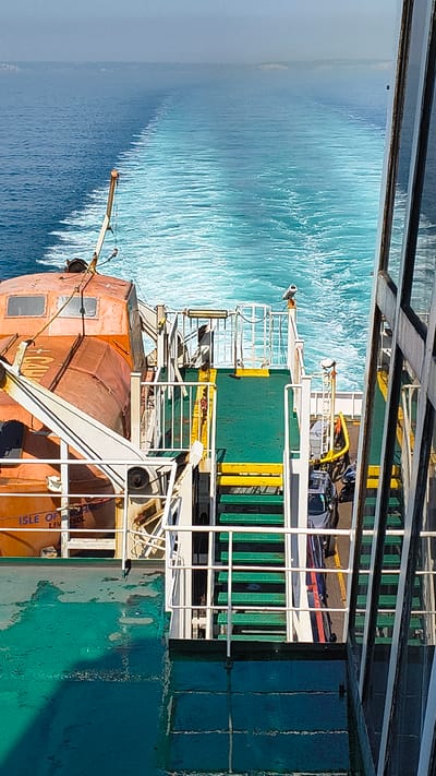 The image shows a view from a ferry deck looking out at the choppy, turquoise sea. The ferry appears to be in motion, as evidenced by the wake trailing behind it. On the deck, various objects are visible, including a bright orange lifeboat, yellow and green barriers, and what appears to be a vehicle secured with straps. White railings and structures of the ferry's upper decks are also prominent.
The lighting suggests it is daytime, with bright sunlight reflecting off the water and the ferry's surfaces. The sea conditions appear to be moderately rough, with whitecaps indicating wind and waves.
Partial text is visible on the side of the orange lifeboat, reading "+ CAV" and "ISLE OF LIM...SOL". The lifeboat is also marked with "ISLE OF MAN SHIPOWNERS" and "LIMERICK" with "LIM" partially obscured. This suggests the lifeboat is associated with a vessel from the Isle of Man.
The overall scene is one of transit and maritime activity. The perspective is from a higher deck of the ferry, looking down and across. No people are clearly visible in the frame. The scene is functional and industrial, typical of a working ferry, but the vibrant blue of the sea adds a natural beauty.