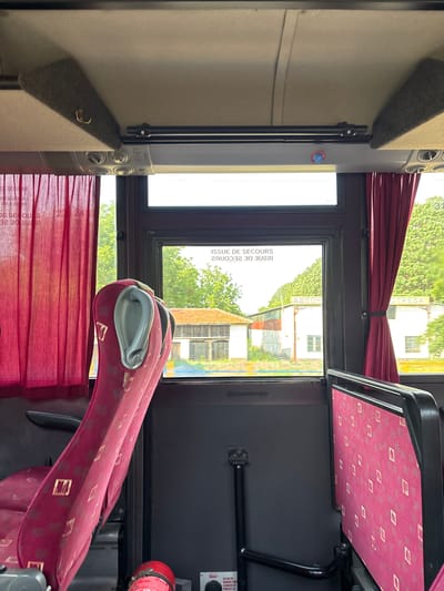 This media file shows the interior of a bus, likely in Malchika, Bulgaria. The scene is captured from a low angle, looking towards the front of the bus and out the window.

The subjects include bus seats upholstered in a red fabric with a repeating diamond pattern in gold. Parts of these seats are visible in the foreground and middle ground. The window area reveals an outdoor scene with green trees and buildings, suggesting daytime with good weather.

Notable details include a red curtain pulled to the side of the window, and text visible on the window itself that reads "ISSUE DE SECOURS" and "DE SECORS" in what appears to be French, possibly indicating an emergency exit or procedure. There are also circular vents visible on a silver panel. A red fire extinguisher is mounted on the wall to the right. The lighting suggests it is daytime.

The overall impression is a functional but somewhat aged interior of public transportation. There are no people visible in the frame. The provided text on the window, along with the mention of "GUVERT" and instructions for danger, points to safety features within the bus.