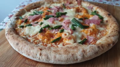 A close-up, overhead shot features a round pizza with a golden-brown, puffy crust. The pizza is topped with melted white cheese, scattered pieces of pink ham, and vibrant green asparagus spears. A creamy orange sauce appears to be the base layer. The pizza rests on a light brown wooden cutting board. In the background, a floral patterned tablecloth with pink and white flowers is visible. The setting is likely indoors, given the lighting and background elements. There are no people or animals present. The image does not provide any specific information or context related to the city of Vrchotovy Janovice, Czechia.