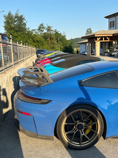 The image shows a line of blue sports cars parked in a row, with a building in the background. The cars are parked on a paved surface, with a metal fence in the foreground. The building appears to be a hotel, with wooden beams and stone accents. Trees and hills are visible in the background, suggesting a rural or mountainous setting. The sky is clear and blue, indicating a sunny day.

The primary subjects are the sports cars, particularly the bright blue one in the foreground which dominates the frame. Its wheel, with a gold rim and yellow brake caliper, is clearly visible. A detail on the car's rear spoiler reads "WEISSACH" in white text with a blue outline. Other cars in the row are also visible, showing different colors, including red and yellow. There is at least one person visible near the building in the background, a dark figure walking.

The setting is outdoors, in what appears to be daylight, likely morning or afternoon given the shadows. The presence of multiple sports cars suggests an event, a gathering, or perhaps a dealership or rental service. The building's architecture hints at a European location, possibly in a resort or tourist area. The overall impression is one of luxury and speed, captured in a bright, clear environment.