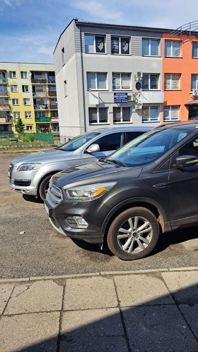 The image shows a street scene in Krosno Odrzańskie, Poland, on a sunny day. Two cars are parked on the right side of the image, with a sidewalk made of concrete slabs to their right. The cars are a silver SUV in the foreground and a dark gray SUV behind it. The SUVs are parked close to each other. In the background, there is a multi-story building with orange and gray facades. The building has many windows, and some of them display signage for businesses, including "CZARNY TATTOO" and "ROTEZOWNIA DENTYSTYCZNE." The building's architecture suggests it is a residential or mixed-use property. The sky is clear and blue, indicating fair weather. The lighting suggests it is daytime, likely mid-morning or afternoon, given the shadows cast by the cars and the building.