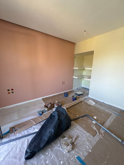 The image displays an interior room located within the Anavissos Municipal Unit, Greece, currently undergoing painting and renovation. The room's ceiling is uniformly white. The left wall is painted a solid terracotta or peach color, while the right wall is light yellow or cream. A recessed, built-in shelving unit with multiple shelves is integrated into the right wall, matching its light yellow/cream coloration.

On the lower section of the terracotta wall, two sets of electrical outlet or conduit openings are visible: four circular openings on the left and three smaller, irregularly shaped openings further right. A single white electrical wire hangs from the ceiling near the junction of the two wall colors. The floor is extensively covered with protective materials, including textured brown cardboard sections and white plastic sheeting, both heavily splattered with white paint. Blue painter's tape secures some of these coverings.

Various painting equipment and supplies are distributed across the floor. Two blue paint cans and a paint roller tray are positioned near the terracotta wall. Closer to the foreground, a small wooden box containing tools or small items, along with several bottles and brushes, rests on a piece of cardboard. A long, gray metallic pole, likely a paint roller extension, lies horizontally across the mid-ground. A large, black plastic bag, conical in shape, occupies the lower center foreground, next to a crumpled, paint-stained white cloth or rag. No individuals are present in the scene.