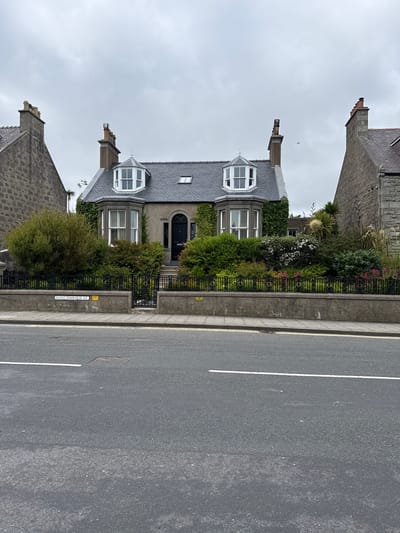 The image depicts a detached house situated on King Harald Street in Lerwick, United Kingdom. The house, characterized by its grey stone facade and slate roof, features prominent bay windows on the front, adorned with white frames. A black front door is centrally located, accessible via a small set of steps. The property is enclosed by a black wrought-iron fence and a low stone wall, behind which a lush garden with various plants and flowers is visible. The street in front of the house is asphalted, with white painted lines indicating a road. The sky is overcast, suggesting cloudy weather. The scene is devoid of any individuals or discernible activity.
