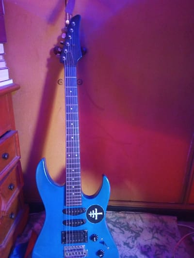 A blue electric guitar stands against a reddish-brown wall. The guitar has a black headstock, a fretboard with dot inlays, and a black and white circular emblem on the body. A wooden dresser with drawers is visible to the left, topped with a stack of books. The image has a purple hue cast by the lighting. A textured rug or cloth lies at the base of the guitar.