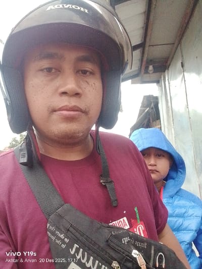 The image depicts two individuals in Jagapura Lor, Indonesia, captured in what appears to be a selfie on December 20, 2025, at 17:17, according to visible metadata from a vivo Y19s device attributed to "Akhter & Arvan". The primary subject, a male, occupies the foreground, looking directly at the camera. He wears a black motorcycle helmet with "HONDA" visible in reverse on the forehead, a maroon short-sleeved t-shirt, and a black fanny pack or sling bag worn across his chest. Stubble is visible on his chin and upper lip, and several small marks are present on his cheeks. A secondary individual is partially visible behind and to the right of the primary subject, wearing a blue hooded jacket with the hood pulled over their head, also looking towards the camera. The background consists of an overhead structure with metal beams, a light-colored wall or partition to the right, and indistinct building structures with visible rooflines, possibly corrugated metal, further in the distance. The lighting suggests daylight conditions, possibly overcast.