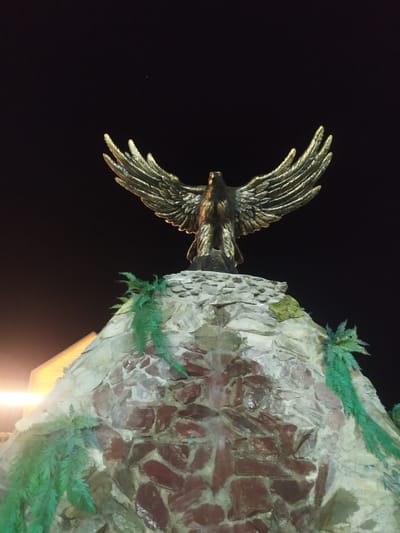 A bronze eagle statue with outstretched wings stands atop a rock formation adorned with artificial green foliage. The statue is illuminated from below, casting a warm glow on its metallic surface. The background is a dark night sky, with a faint light source visible on the left side of the image, suggesting artificial lighting. The rock formation appears to be constructed with irregular stones of varying colors, predominantly shades of brown and gray, and is textured to resemble a natural cliff face. This monument is located in the city of Sosnovka, Kyrgyzstan.