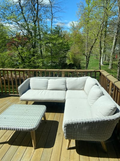A white wicker sectional sofa with white cushions sits on a wooden deck overlooking a grassy backyard. There is a small wicker table in front of the sofa. The backyard is filled with lush green trees, some of which are deciduous and some evergreen. The deck is surrounded by a wooden railing. The scene is located in Rye, New York, United States (latitude: 40.952, longitude: -73.708). The sun is shining brightly, casting shadows on the deck and the furniture. The scene is calm and peaceful.