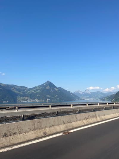 The image depicts an elevated, panoramic view of a Swiss landscape, consistent with a vantage point from the A2 highway near Beckenried, Switzerland. In the foreground, a dark asphalt road surface with a distinct white edge line diagonally recedes from the lower right. Adjacent to the road is a concrete highway barrier exhibiting signs of wear and discoloration, topped by a metal guardrail system featuring a lower mesh infill section. A rectangular metal grate is visible embedded in the asphalt on the lower right. Beyond the barrier, a calm body of water, identified as Lake Lucerne, spans the midground horizontally. Along the opposite shore of the lake, the town of Beckenried is visible as a collection of structures and buildings. The background features a series of prominent mountains; the most striking is a large, pointed, and heavily forested peak, likely Buochserhorn, rising directly from the lake. Further in the distance, additional rugged mountains with some visible rock faces and subtle snow patches are present. The sky above is clear and uniformly blue, with minimal, wispy white clouds near the horizon on the right. No individuals, vehicles, or dynamic actions are depicted.