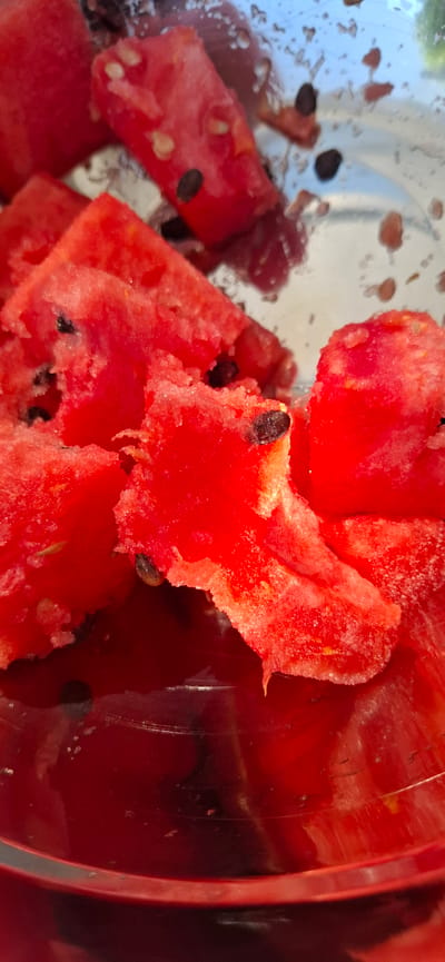 The image shows a close-up of chopped watermelon pieces with black seeds inside a clear container, likely a bowl or glass. The watermelon is a vibrant red, and some pieces appear to be slightly juicy or wet, with small droplets visible. The container itself has a reflective surface, showing some light glare and indistinct shapes in the background. The focus is on the texture and color of the watermelon. There is no indication of the city of Nörvenich, Germany, or any specific location within a city. The scene is static and focuses solely on the food item.