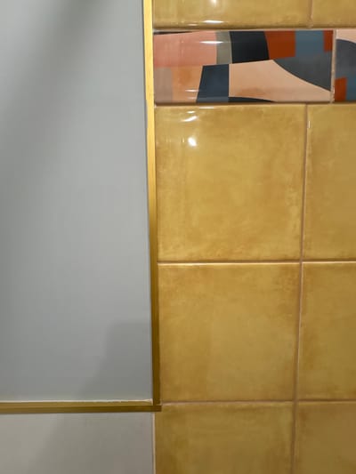 The image shows a close-up view of a wall in a building located in Dolce Vita KP, Russia. The wall is divided into two distinct sections by a thin, gold-colored trim. The left section of the wall is a solid, light gray color, possibly painted. The right section is covered in square, glossy, mustard-yellow tiles. Above the yellow tiles, there is a horizontal band of rectangular tiles with a geometric pattern featuring shades of peach, dark blue, rust, and light gray. The lighting in the scene is bright, creating highlights on the glossy surfaces of the tiles. The photograph is taken from a slightly elevated angle, focusing on the texture and color of the wall finishes. There are no people or actions depicted in the image.