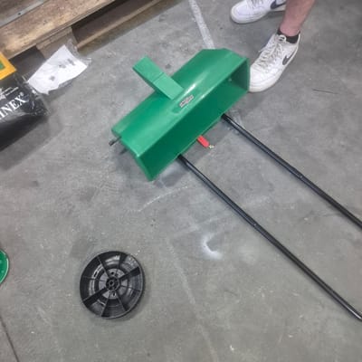 The image shows a green seed spreader lying on a concrete floor, possibly in an industrial setting, as suggested by a wooden pallet in the background and a bag labeled "LINEX". Two black metal poles extend from the seed spreader, suggesting it is designed for walking or pushing. To the left of the spreader is a black plastic wheel with a spoke design. In the background, a person's feet wearing white Nike sneakers are visible. The overall scene is consistent with a storage area or workshop. The image does not provide any information about the city of Nowa Wieś Książęca, Poland.