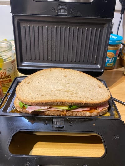 An unpressed sandwich rests on the open bottom plate of a black electric sandwich maker or grill, positioned on a light brown wooden countertop. The sandwich consists of two slices of light brown, textured bread. Between the bread are visible layers: a reddish-orange spread, pink deli slices (likely ham), light green vegetable slices (possibly cucumber or zucchini), and dark green leafy vegetables (possibly spinach or rocket).
The sandwich maker's top plate is elevated, revealing its clean, ribbed, non-stick grilling surface. The bottom plate exhibits minor orange-yellow residue around the sandwich. A black handle mechanism is visible on the upper part of the appliance.
In the background, on the left, a clear glass jar with a yellow label displaying French text ("SANDWICH", "CORNICHONS", "TRANCHES") contains pickled items. To the right, a partially visible blue-capped jar with "rs!" and "CREMEUX" on its label contains a creamy substance, consistent with peanut butter. A black electrical cord extends upwards from behind the sandwich maker on the right side. The background wall is light-colored.
The image itself provides no discernible visual cues to pinpoint a precise location within Boulogne-Billancourt, France, beyond the presence of French language labels consistent with the country.