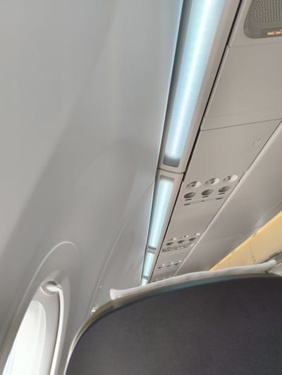 The image shows the interior of an airplane cabin. The perspective is looking upwards towards the overhead compartments and lighting. A headrest of a passenger seat is visible in the foreground, with a white pillow placed on it. Above, there are rows of illuminated light strips and ventilation controls with circular vents. The overall color palette is dominated by white and grey, typical of an aircraft interior. The image does not contain any information to determine a specific location in Thái Hưng, Vietnam.