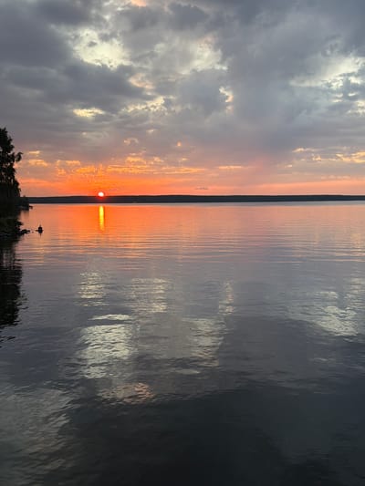 This is a vertical, wide-angle photograph of a sunset over a body of water. The sun, a bright orange orb, is low on the horizon, casting a vibrant orange and yellow glow on the clouds and water. The sky on the left is filled with dramatic, grey clouds interspersed with patches of bright sunlight. On the right, the water is dark and reflective, mirroring the colors of the sky and the sun's light. A dark line on the horizon suggests a distant shore or landmass. In the foreground, the dark silhouette of trees frames the bottom left of the image. A small, dark boat is visible on the water, near the center. The overall mood is serene and peaceful, with the dramatic colors of the sunset creating a sense of awe. The time of day is clearly sunset. The weather appears to be partly cloudy. While no specific location cues are visible, the description provided indicates the location is Zarechny, Russia. There is no visible text in the image.