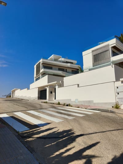 The image depicts a modern, multi-story white building with a flat roof and glass balconies, situated on a slight incline. The architecture features clean lines and geometric shapes, with some sections having vertical slat detailing. A high white wall with a dark, retractable gate encloses the property. The foreground shows a pedestrian crossing with white stripes painted on a paved road. The sky is a clear, bright blue. The scene appears to be in a sunny, urban or suburban environment, likely in Dehesa De Campoamor, Spain, given the architectural style and clear skies commonly associated with the region. There are no people or animals visible in the image. The overall impression is one of contemporary, luxurious residential property bathed in sunlight.