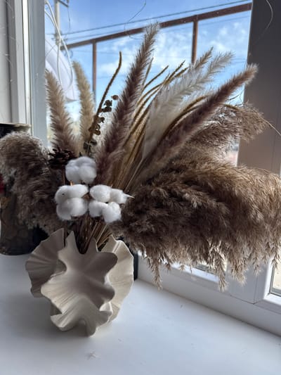 The image, oriented vertically, captures a close-up still life of a dried floral arrangement. The bouquet is positioned towards the bottom and center, with a window and outdoor scene visible on the left, and a white wall or windowsill extending to the right.

The arrangement prominently features voluminous plumes of pampas grass in various natural shades of beige, brown, and off-white, giving a soft, feathery appearance. Tucked among the grass are several pristine white cotton bolls, a small pinecone, and other slender dried botanical elements, some with tiny seed structures.

To the upper right of the bouquet, a distinctive light-colored decorative object rests on a white surface, possibly a windowsill. This object has an organic, ruffled form, resembling overlapping leaves or petals, and appears to be made of ceramic or a similar textured material. A dark container, likely a vase, is partially visible at the bottom, holding the bouquet.

Through the window on the left, a clear blue sky with sparse white clouds is visible, indicating bright daytime weather. The window frame shows distinct divisions. The scene conveys a calm, natural, and earthy aesthetic, highlighting the beauty of dried flora. No activity is depicted, and no text is visible. While the provided context notes the location as Semenovka, Kyrgyzstan, this information is not discernible from the visual cues within the image.