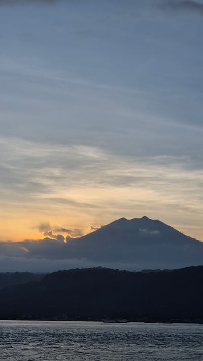 The image depicts a landscape at sunset, possibly in Bulusan, Indonesia. A dark, silhouetted mountain range dominates the right side of the frame, bordering what appears to be a body of water. To the left of the mountain, the sky transitions from light blue to golden orange near the horizon, with wispy clouds reflecting the sunset colors. A few bright clouds are illuminated along the mountainside.