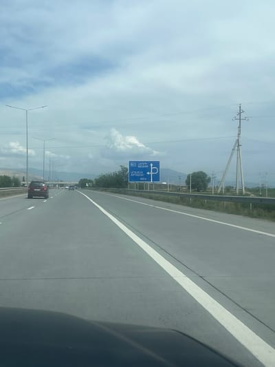 The image captures a daytime view from inside a vehicle traveling on a multi-lane highway in Armenia, near Ayntap. The sky is bright with scattered clouds, indicating fair weather.

On the left side of the highway, a prominent blue road sign provides navigational information. The sign features a U-turn arrow symbol and lists destinations in Armenian script and Latin transliteration: "M.2", "ՄԵՂՐԻ" (Meghri), "ՍՈՒՍՈՒՍ" (Susus), and "ԱՐՏԱՇԱՏ" (Artashat), along with "400 M," indicating the distance to these points or the upcoming turn. Beyond the sign, large power line structures with multiple cables run parallel to the road. A metal guardrail separates the highway from the adjacent terrain, where sparse vegetation and a single prominent tree are visible.

The highway itself is asphalt with clear white lane markings. Several vehicles are seen ahead on the road, with a red SUV being the most distinct in the middle distance. In the far background, hazy mountains stretch across the horizon. The right side of the frame shows a partial view of the interior of the vehicle, likely a window frame or dashboard element. The scene depicts typical highway travel.