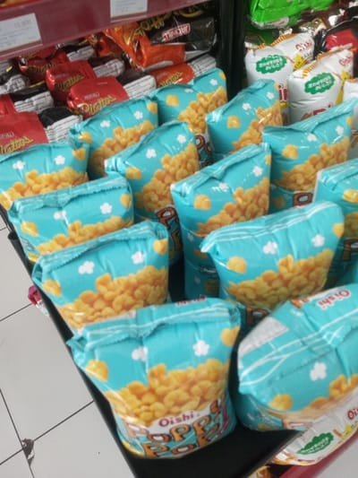 This image depicts the interior of a retail store in Naung Bo Oe, Myanmar. The scene is dominated by a display of packaged snack foods. In the foreground and midground, numerous bags of "Oishi Poppo Pop" snacks are arranged on a dark-colored shelf. These bags are primarily turquoise with a clear window revealing light-yellow, irregularly shaped puffed snacks. The turquoise portion features white cloud-like designs and small yellow paw prints. The brand name "Oishi Poppo Pop" is printed in prominent red and blue lettering at the bottom of the bags. Behind these, on higher shelves, various other snack packages are visible, including red bags labeled "Platitos," orange bags with partially visible "Jack N Jill" branding, and stacks of green and white "Oishi" branded bags, some with foreign language text. A red price tag displaying "10.900" is attached to an upper shelf on the left. The floor in the lower left corner is a light-colored tile with dark grout lines. No individuals are present, and no active interactions are occurring within the frame.