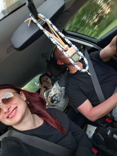 Captured from inside a moving vehicle, the image shows three individuals and various objects in Любанскі сельскі Савет, Belarus.
The foremost person, on the left, is a woman wearing sunglasses, a black shirt, and a seatbelt. Her hair is dyed a deep red. In the back seat, another woman is visible. She has a clear plastic bag next to her. In the driver's seat is a man also wearing a black shirt and a seatbelt.
Hanging from the rearview mirror is a beaded car accessory, adorned with eye-shaped charms and a perfume tag that reads "TOM FORD TABACCO VANILLE." The vehicle's interior is visible, including the dashboard and door panel. Outside the car, blurred trees suggest the car is in motion.