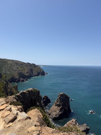 This image depicts a scenic coastal view from a high vantage point overlooking the Atlantic Ocean. The foreground features rugged, rocky cliffs with sparse vegetation. The mid-ground shows a dramatic coastline with steep cliffs plunging into the ocean, where waves crash against the rocks. The ocean itself is a deep blue-green, stretching out to the horizon. In the distance, a faint line of land can be seen, possibly hinting at further coastline or islands. The sky is a clear, bright blue, indicating a sunny day.

This scene is characteristic of the Cabo da Roca area, which is located near the village of Colares in Portugal. Cabo da Roca is known as the westernmost point of mainland Europe and offers spectacular cliffside views of the Atlantic.