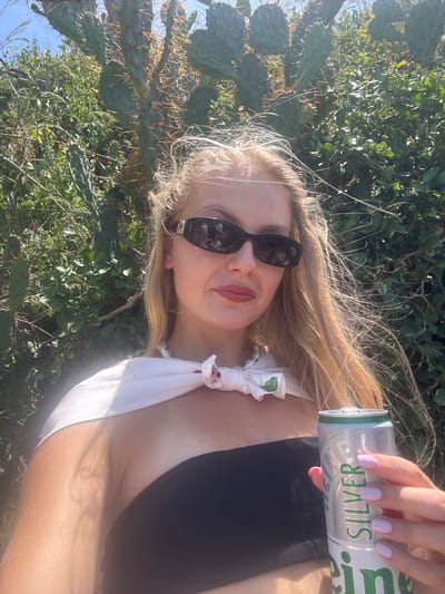 A woman with blonde hair and sunglasses is holding a silver can in her hand. She is wearing a black top and a white scarf with a floral pattern tied around her neck. Her fingernails are painted a light purple. The background is a natural setting with green bushes and cacti. The lighting suggests it is daytime, likely outdoors. The woman appears to be taking a selfie. The can has the word "SILVER" visible on it in green text.
