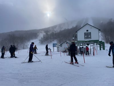 This image captures a bustling scene at the base of a ski lift on a snowy, overcast day in Jay, United States. Numerous individuals, clad in winter ski and snowboard gear including helmets and goggles, are gathered on the snow-covered slope. Some are standing in what appears to be a queue, guided by red ropes, preparing to board a ski lift. Others are already entering the lift loading area.
In the mid-ground, a prominent white building with a dark green roof houses the mechanics for the ski lift, which is actively loading passengers. To the left of this main lift building, a smaller structure is visible, potentially a control hut or service building. The ground is covered in fresh snow, showing tracks from skis and snowboards.
The background reveals a snow-covered mountain, its peaks and upper slopes partially obscured by thick fog and clouds, adding to the atmospheric conditions. Patches of trees, barren of leaves due to winter, dot the mountainside. Hints of groomed ski trails can be seen ascending the distant slope. The sky is largely overcast, but a bright, hazy sun is faintly visible breaking through the clouds in the upper left quadrant, casting a soft, diffuse light on the scene. Snowflakes are visibly falling throughout the image, indicating ongoing light snowfall. No specific text on signs is clearly legible. The overall impression is one of a typical winter day at a busy ski resort.
