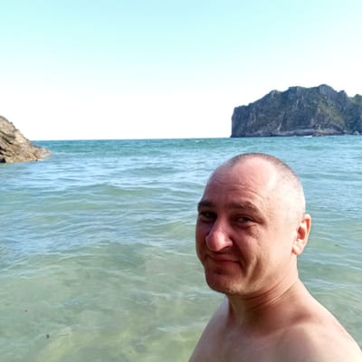A man is taking a selfie in the ocean with an island in the background. He is facing the camera and smiling. The water is clear and blue, and the sky is a bright, cloudless blue. The man is shirtless and appears to be in good health. The island is rocky and covered in greenery. The location is the coast of Ribadedeva, Spain.
