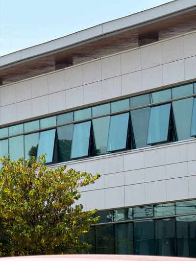 The image shows the exterior of a modern building with a facade of white panels and large windows. Some of the windows are open at an angle, creating a geometric pattern. A tree with green and yellow leaves is visible on the right side of the image, partially obscuring the building. The sky is light blue, suggesting it is daytime. The lighting indicates it may be morning or late afternoon, with sunlight casting shadows and highlighting the foliage. The building's architecture suggests an urban or business park setting. There is no visible text in the image. The scene is static and devoid of any human activity. The overall impression is one of architectural design and the interaction of the built environment with nature. The location context provided is Rungis, France, which may suggest this is a commercial or administrative building within that area.