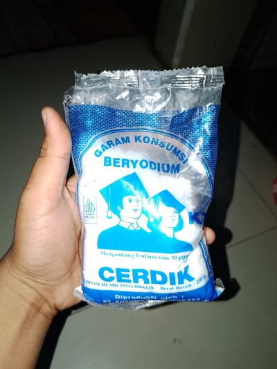 A human hand, with visible skin texture and fingernails, is holding a sealed, transparent plastic package of iodized salt. The packaging is primarily blue and white, featuring text and graphics. Prominently displayed is the brand name "CERDIK" in large blue letters. Above this, in a curved blue font, is "GARAM KONSUMSI BERYODIUM." A graphic of two figures wearing graduation caps is visible on the front. A "HALAL" certification logo is present on the left side of the package. The packaging also indicates "Mengandung Yodium min 30 ppm" and "Berat Bersih : 250 gr". The background is dark and out of focus, suggesting an indoor setting. The image was taken in Sendangrejo, Indonesia.