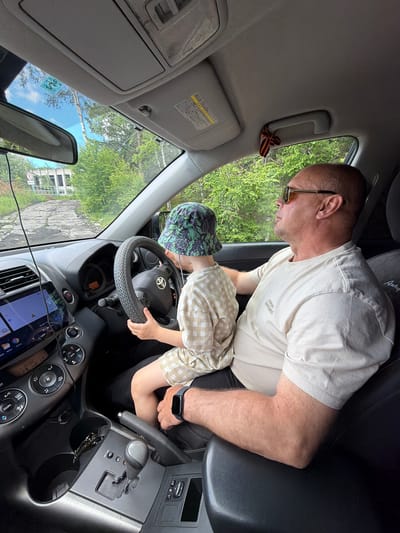 An adult male wearing sunglasses and a light-colored t-shirt sits in the driver's seat of a car, with a young child wearing a bucket hat and a checkered shirt sitting on his lap and holding the steering wheel. The car is on a paved road in what appears to be a rural or semi-rural area, with trees and some dilapidated buildings visible in the background. A "stop" sign is visible on the left side of the road. The interior of the car shows the dashboard, steering wheel, gear shift, and a touch screen display. A ribbon with orange and black stripes is tied to the car's ceiling handle. The scene appears to be set in the city of Kul'dur, Russia, as indicated by contextual information.
