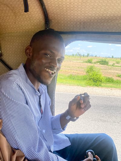 In Yali, Nigeria, a man wearing a blue and white striped shirt and dark pants is seated inside a vehicle. He is smiling and looking towards the camera. In his right hand, he holds a set of keys. The background shows a dry, grassy landscape with scattered trees under a bright sky with clouds. The interior of the vehicle is lined with beige fabric.