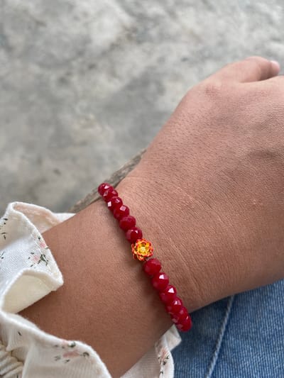 A close-up shot shows a person's wrist adorned with a red beaded bracelet featuring a golden floral charm. The wrist is against a textured grey surface, possibly concrete or stone. The person is wearing a white blouse with a floral pattern and blue denim jeans. The scene is likely outdoors, given the natural lighting and background texture. The image does not contain any information about a specific location in Kutalhoksukon, Indonesia.