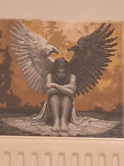 The image displays a tapestry or cross-stitch artwork depicting a person with large wings. The person is seated with their arms crossed, appearing to be in a fetal position. One wing is white, and the other is black, symbolizing duality. The background is a gradient of orange and brown, suggesting a sunset or abstract landscape. The artwork is framed and hangs against a wall, with a radiator visible at the bottom of the image. There is no information within the image to indicate a specific location in Mińsk Mazowiecki, Poland.