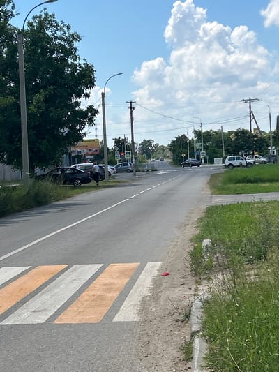 The image depicts a rural roadside scene in Dondukovskaya, Russia, on a clear, sunny day. The perspective is from the side of a paved road, with a grassy embankment to the right. Several cars are parked along the side of the road, interspersed with trees and utility poles. A pedestrian crossing with diagonal white and orange stripes is visible on the asphalt in the foreground, indicating a road crossing.
In the distance, more cars and roadside infrastructure, including traffic lights and street lamps, are visible. The sky is bright blue with scattered white, puffy clouds, suggesting it is daytime, likely late morning or early afternoon. The presence of lush green grass and trees indicates a warm season, possibly summer or late spring.
There are no distinct human activities happening, with the focus being on the parked vehicles and the general environment. One car in the middle distance has a person leaning into it, but their activity is unclear. The overall atmosphere is one of stillness and a typical day in a less urbanized area. Some faint text is visible on a yellow sign in the distance, but it is illegible.