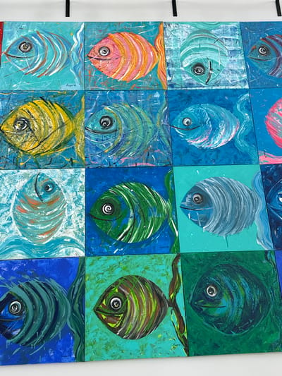 The image shows a wall decoration composed of twelve square paintings of fish. Each fish is painted in a different color and style, but all face the viewer. The background of each painting varies in color and texture. The paintings are arranged in a 3x4 grid. The location is Kalyves Polygyrou, Greece.
