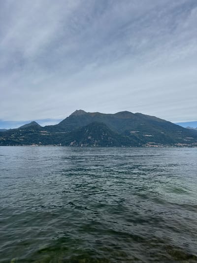 This image captures a scenic view of Lake Como near Gittana, Italy. The foreground is dominated by the calm, dark blue water of the lake, with ripples reflecting the overcast sky. In the middle ground, a lush, green mountainside rises steeply, dotted with small clusters of houses and buildings that form a village along the slopes. The peaks of the mountains are partially obscured by a cloudy sky. The overall impression is one of natural beauty and serene Italian landscape.