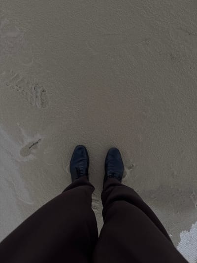 The image displays a downward, overhead perspective of a person's lower legs and feet on a sandy beach in Grottammare, Italy. The individual is wearing dark brown trousers and dark, lace-up shoes. The footwear is positioned on fine-grained, light brown to grey sand. The sand appears partially damp, notably around the shoes and in the foreground towards the lower right, indicating proximity to the waterline or a recently wet environment. Faint, irregular depressions and subtle ripple patterns are visible in the sand, consistent with prior foot traffic or natural water action. No other individuals or significant objects are present within the frame.