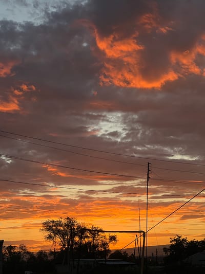 The image captures a dramatic sunset over Nor Kharberd, Armenia. The sky is filled with a gradient of colors, from deep oranges and fiery reds in the lower half to dusky purples and grays higher up. Wispy clouds are illuminated by the setting sun, creating a textured and dynamic celestial canvas. Silhouetted against this vibrant sky are the dark shapes of trees, power lines, and the outlines of buildings and a utility pole in the foreground. The overall impression is one of a serene yet powerful natural display, framed by the elements of human habitation and infrastructure.