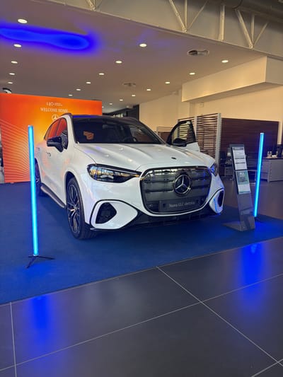 The image shows a white Mercedes-Benz SUV on display in a showroom, likely a car dealership, located in Parma, Italy. The car is positioned on a dark gray tiled floor, with a bright blue carpet area in front of it, illuminated by blue LED lights emanating from vertical light stands. Behind the car, an orange backdrop with text that appears to read "140 YEARS OF INNOVATION WELCOME HOME" is visible. The car's front doors are open, revealing part of the interior. To the right of the car, a display stand holds information brochures, including one with the text "ALL EXCLUSIVE". The overall environment is clean and well-lit, with ceiling lights and ambient blue lighting contributing to the modern showroom atmosphere.