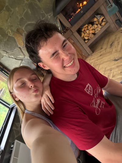 In a dimly lit room with a stone wall and a fireplace, a young man and woman are taking a selfie. The man, with dark hair, is smiling and winking at the camera. The woman, with blonde hair, is puckering her lips as if for a kiss, with her eyes closed. She has her arm around the man's neck. The fireplace in the background has a fire burning and logs stacked next to it. The floor is made of wooden planks. A large window on the left side of the image shows a glimpse of greenery outside. A white air conditioning unit is visible on the wall near the window.