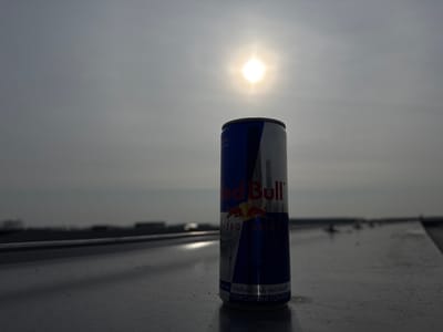 A standard Red Bull energy drink can, predominantly blue and silver with the brand logo and "ENERGY DRINK" text visible, stands upright on a flat, light-colored surface in Naaldwijk, Netherlands. The can is positioned slightly right of the frame's center. The background consists of a hazy, light-gray sky with the sun clearly visible as a bright, starburst-like point in the upper-left quadrant, backlighting the scene. A blurred, dark horizon line featuring indistinct distant structures or vegetation is present below the sun. No individuals, actions, or interactions are depicted.