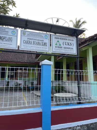 The image shows a signpost with multiple signs in front of a school building. The signs are for "PEMERINTAH KABUPATEN BOYOLALI DINAS PENDIDIKAN DAN KEBUDAYAAN" (Boyolali Regency Government, Department of Education and Culture), "PERPUSTAKAAN CAKRA BUANA ILMU" (Cakra Buana Ilmu Library), and "USAHA KESEHATAN SEKOLAH (UKS) SD NEGERI 1 KEDUNGPILANG" (School Health Business, State Elementary School 1 Kedungpilang). The building appears to be an elementary school with green walls and a tiled roof. A white metal fence is in the foreground, with blue and red painted posts. In the background, there are trees and a satellite dish. The location is identified as Kedungpilang, Kecamatan Wonosamodro, Kabupaten Boyolali, Indonesia. The overall scene depicts the entrance and signage of an educational institution.