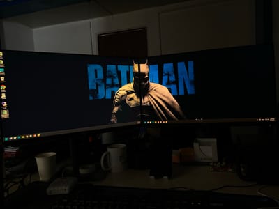 The image depicts a dimly lit office desk with a large, curved monitor displaying a "BATMAN" logo and a promotional image of the Batman character. The monitor is positioned in front of a wall with what appears to be a doorway. On the desk in front of the monitor, there are several objects including a white paper cup, a white mug with a logo, a smartphone in a stand, a notepad holder with sticky notes, and a black keyboard. The overall impression is one of a workspace at night or in low light conditions. The image does not contain any information that would allow for the determination of its location in Yukhta, Russia.