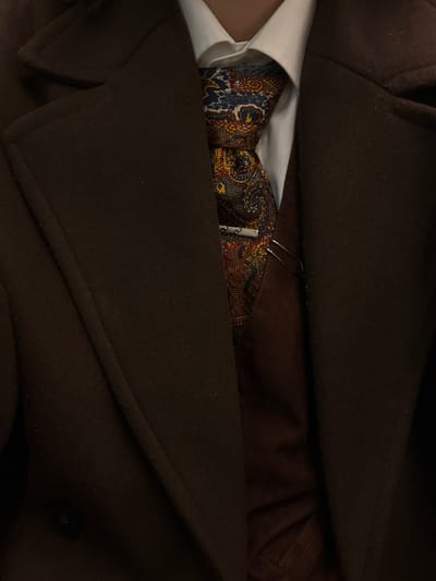 A dark brown overcoat is seen open, revealing a white collared shirt and a patterned tie with a tie clip. The tie features intricate designs in gold, blue, and red hues. A silver tie clip is visible. The inner lining of the coat appears to be brown. The scene is captured in subdued lighting.