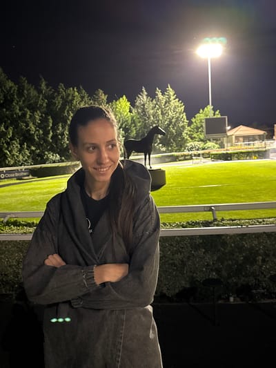 The image captures a woman standing in an outdoor setting, likely at night. She is positioned to the right of the frame, dressed in a dark-colored hoodie and jeans, and smiling at the camera.
Behind her, the scene is illuminated by artificial lighting. There is a grassy field, a horse statue, along with trees and bushes. A building is visible in the background. The presence of a sign that says "Close Brothers" suggests the location is a sports venue, possibly a racecourse.
Given the prompt's specifications, it is likely that this scene is located in the Borough of Spelthorne, United Kingdom.