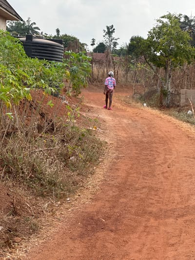 The image depicts an outdoor scene in Agbogugu, Nigeria, featuring a reddish-brown dirt road under an overcast sky. A single individual, observed from behind, walks away from the camera along the road. This person wears a light-colored head covering, a pink short-sleeved t-shirt with a blue and white graphic on the back, patterned shorts or a skirt, and pink slip-on sandals. To the left of the road, an embankment of red earth supports dense green-leafed plants, including large-leafed vegetation, and smaller dry bushes with white flowers. Partially obscured by this foliage, a black horizontal cylindrical water tank is visible, resting on a structural base. A section of a light-colored building with a dark roof is present in the upper left background. To the right of the road, a small tree with green foliage stands, and further back, stacked concrete blocks are visible. The overall background consists of sparse trees, dry vegetation, and distant structural outlines.
