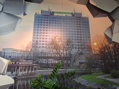 The image depicts a mural or large-scale print of a cityscape, possibly in or representing the city of Piła, Poland. The scene is dominated by a large, modern hotel building with a prominent green sign reading "HOTEL GROMADA" at the top. The hotel is multi-story with numerous windows. In the foreground, a body of water with reflections is visible, and on the water, there is a long, multi-level boat or floating structure with a red roof. Trees, some bare and some with leaves, are scattered throughout the scene, suggesting either late autumn or early spring. The sky has a warm, sunset or sunrise glow. The image also includes abstract, geometric shapes overlapping the top and sides, suggesting it might be displayed indoors or as part of an artistic installation. There are no people visible in the depicted scene.