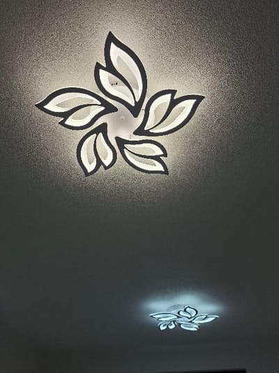 The image displays an interior scene from Krupnik, Bulgaria, featuring two illuminated LED ceiling light fixtures against a textured grey ceiling. The primary fixture, positioned towards the top and center, is a large, multi-component unit designed in a stylized floral or leaf pattern, with bright white translucent sections outlined in dark grey or black. This fixture emits a strong white light, casting illumination onto the immediate surrounding ceiling surface, which exhibits a fine, granular texture. A secondary, smaller light fixture of similar design, less brightly illuminated and appearing further away, is visible in the lower right quadrant of the image. No individuals, objects, actions, or interactions are present in the frame beyond the ceiling and light fixtures.