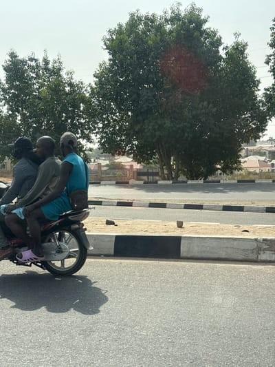 An outdoor scene in Aso Housing Estate, Nigeria, captured in bright daylight. Three adult males are closely seated on a dark motorcycle, positioned left of a concrete median strip. The driver is partially obscured, wearing a dark top and dark cap. The middle passenger wears a light-colored long-sleeved shirt and blue shorts. The rearmost passenger wears a light blue sleeveless top, light blue shorts, a light-colored cap reversed on his head, and pink slide sandals; a dark satchel is slung across his shoulder. The motorcycle is on an asphalt road. The median strip features alternating black and white painted sections and a sandy area containing two small, dark irregular objects. In the background, large green-leafed trees stand before a residential area with buildings featuring light-colored roofs and a dark perimeter fence. A hazy sky is visible above. Strong shadows from the motorcycle and riders are cast on the road.